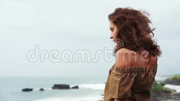 可爱的成熟女人棕色卷发站在悬崖边凝视海浪视频的预览图