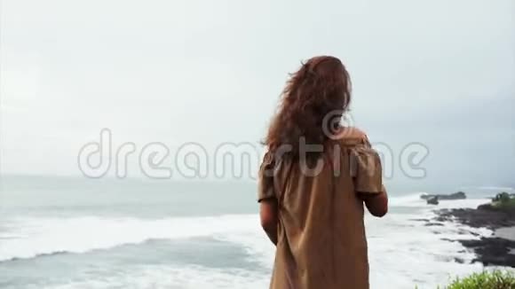 漂亮的女人棕色卷发站在悬崖边凝视着海浪视频的预览图