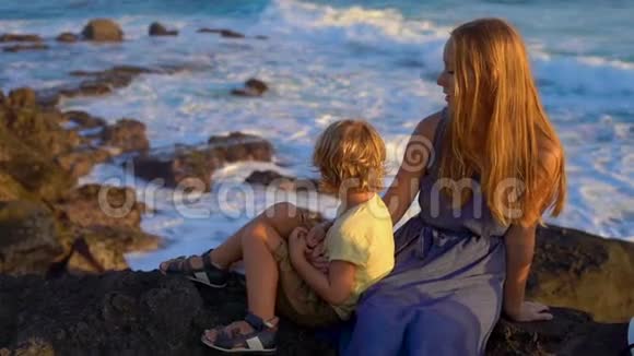 一对母亲和儿子坐在岩石上看着塔纳罗得寺附近的海浪慢镜头视频的预览图