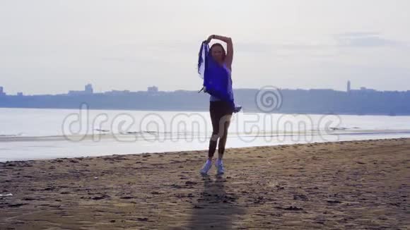 穿着运动服带着蓝色围巾跳舞的年轻苗条黑发女人一个女人在一个大丽的沙滩上订婚视频的预览图