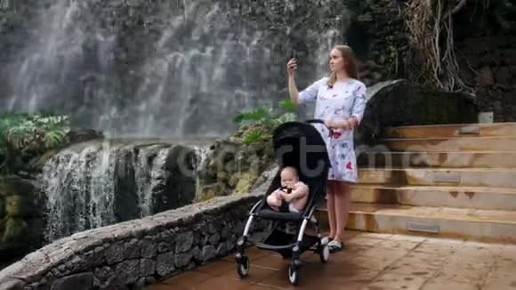 一位年轻的母亲带着她的孩子在瀑布附近散步在她的智能手机上与家人一起旅行时拍照视频的预览图