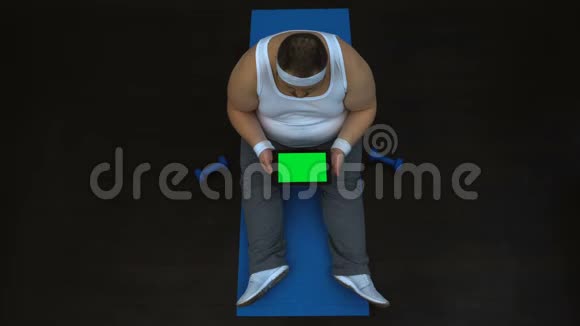 肥胖男性滚动式平板电脑坐健身房阅读健康生活方式文章视频的预览图