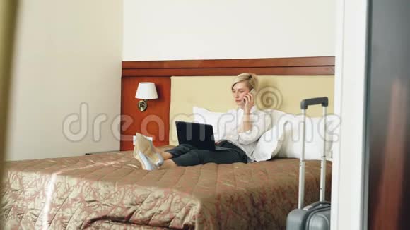 酒店客房内穿着白色衬衫的女商人在床上躺着用笔记本电脑和手机聊天视频的预览图
