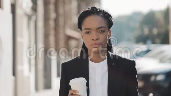 一位严肃的非洲裔美国女商人穿着西装在城市里走来走去喝着咖啡看着视频的预览图