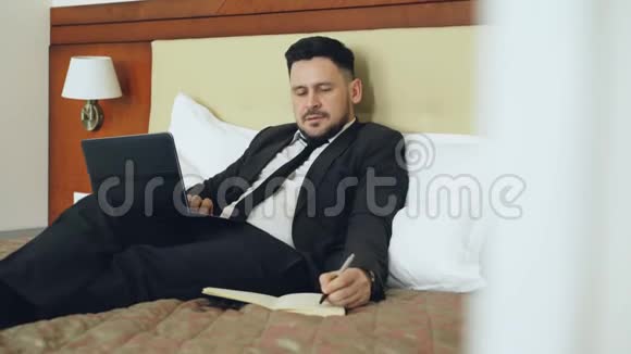 微笑的商人躺在床上使用笔记本电脑和在记事本写笔记同时在酒店房间工作旅费视频的预览图