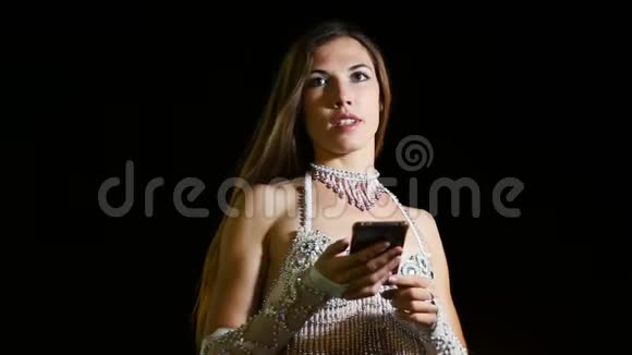 穿着东方民族服装的年轻女舞者晚上在户外使用智能手机视频的预览图