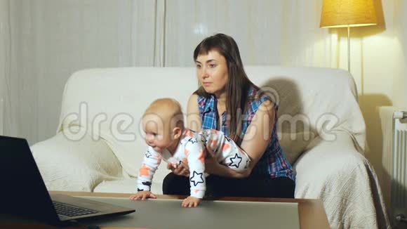 妈妈和蓝眼睛宝宝坐在白色沙发上看笔记本电脑屏幕上的视频视频的预览图