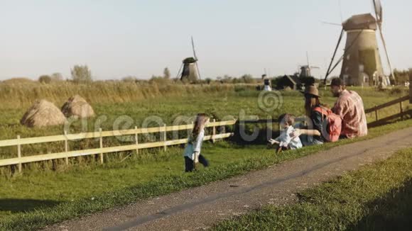 一家四口坐在风车农场附近妈妈爸爸和两个孩子欣赏阳光明媚的夕阳乡村风光4K视频的预览图
