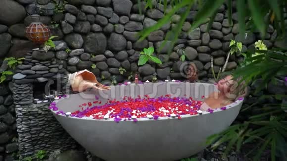 美丽的模特躺在充满玫瑰的室外浴缸里的水中水疗院铺有石头装饰精美视频的预览图
