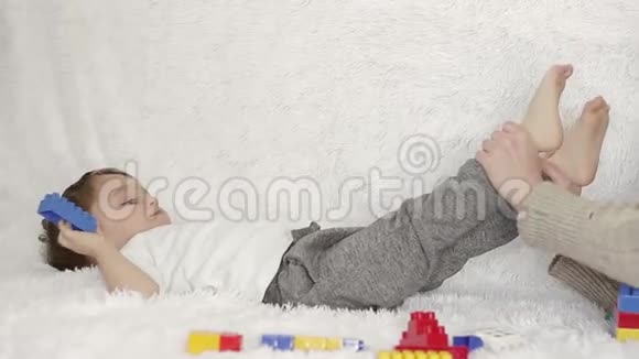一位快乐的母亲亲吻孩子双腿躺在白色沙发上背景是彩色玩具视频的预览图