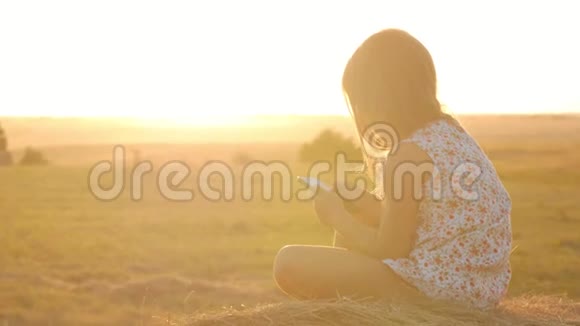 日落时分小女孩用手机坐在收获卷上漂亮的少女在干草堆上视频的预览图
