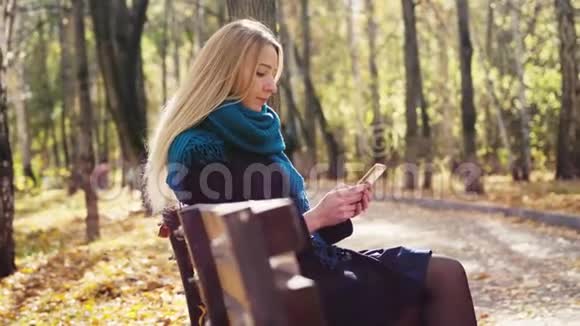秋天公园里坐在长凳上用智能手机的金发美女3840x21604公里视频的预览图