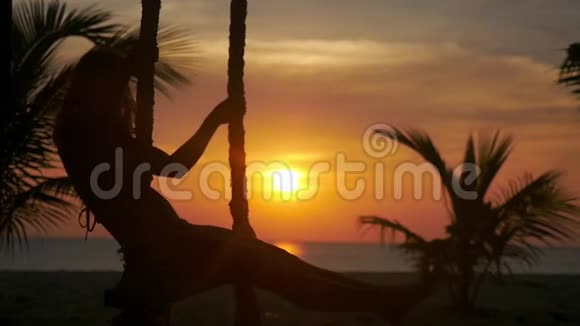 美丽的年轻女子在令人惊叹的夕阳下在热带海滩的秋千上摇摆慢动作1920x1080视频的预览图