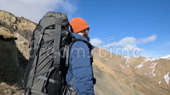 一个长胡子的人戴着太阳镜帽子背着背包和照相机站在山上看着场边视频的预览图