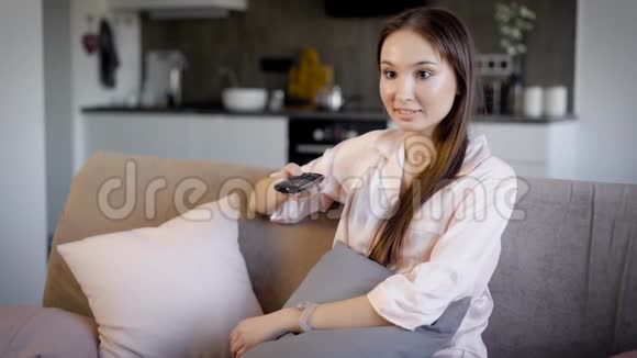 漂亮的黑发女人在度假时躺在客厅的沙发上通过遥控器切换电视频道视频的预览图