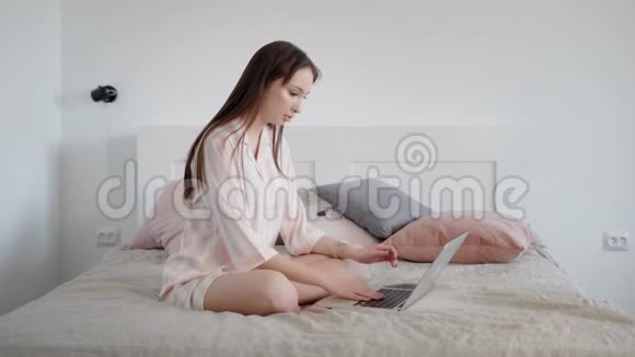 年轻放松的黑发女人白天坐在她家房间的床上在笔记本电脑上打字视频的预览图