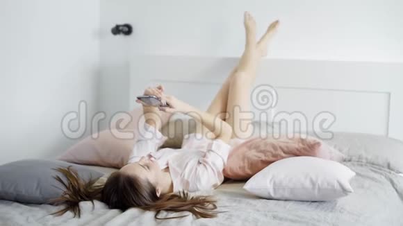 无忧无虑的年轻黑发女人躺在床上翘着双腿站在床头板上用互联网输入信息和张贴信息视频的预览图