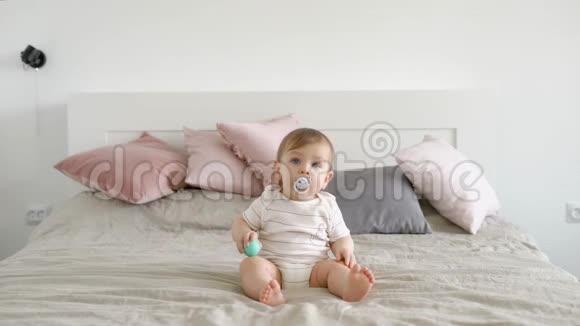 金发碧眼的婴儿坐在一张大床上带着婴儿奶嘴白天在舒适的家中向前看视频的预览图