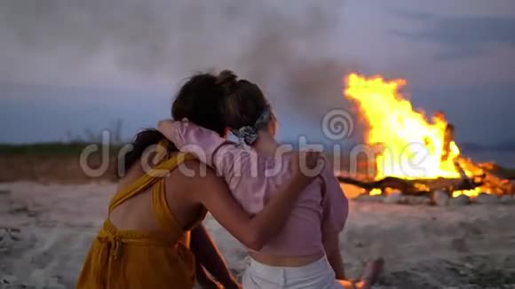 两个女朋友拥抱在火前的原木上回到镜头前体贴地注视着火中的火焰视频的预览图