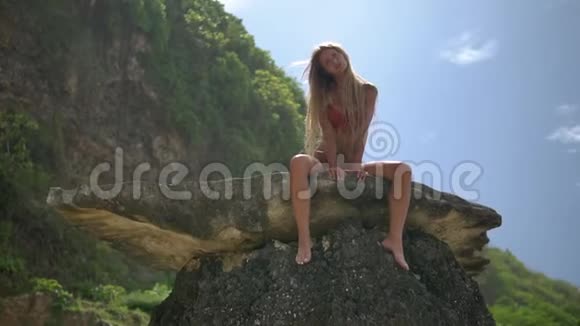 可爱的成年女孩穿着蓝色比基尼双脚悬在丛林中间的悬崖上长长的金发遮住了她的身体视频的预览图