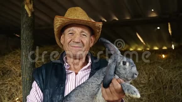 一个戴草帽的老农夫正抱着一只灰色的大兔子干草背景下男人的肖像视频的预览图