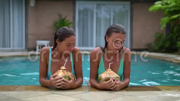 两个穿着蓝色粉末比基尼的可爱的成年女孩在室外游泳池里用吸管喝椰子鸡尾酒视频的预览图