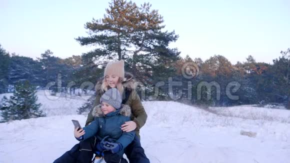 冬天的家庭带着手机的小家伙在雪山上的雪橇上和妈妈一起拍视频视频的预览图
