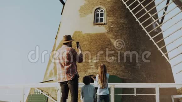 有两个孩子在乡村风车附近的男人带小孩的爸爸在他的智能手机上拍摄了一段现场视频后景4K视频的预览图