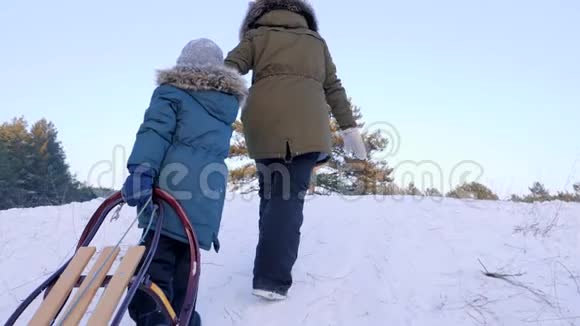 冬天的娱乐活动妈妈和儿子手里拿着雪橇爬上白雪覆盖的小山视频的预览图
