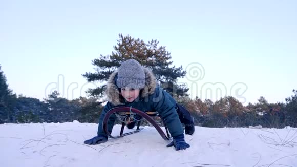 快乐的男孩举起双手躺在雪橇上慢慢地从冬天的森林里的雪山上骑过去视频的预览图