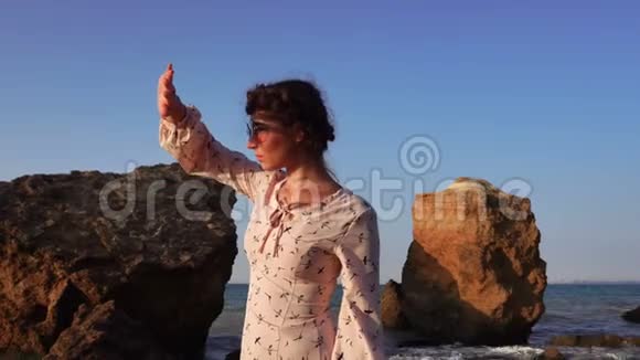 日出时可爱的白种人深褐色的女孩戴着太阳镜和浅粉色的连衣裙坐在石头上视频的预览图