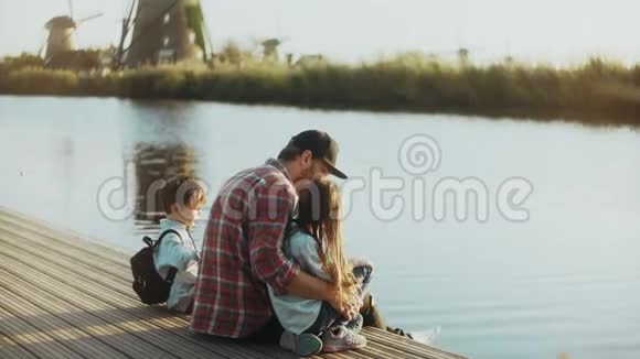 有两个孩子的人坐在小湖码头上戴着黑色棒球帽的单身爸爸家人把时间花在户外4K视频的预览图
