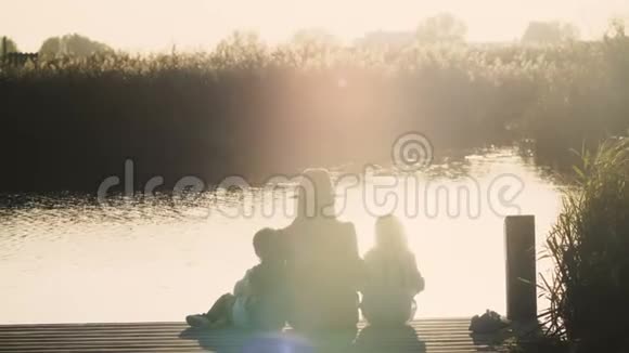 日出时母亲和两个孩子坐在湖边码头上和平与和谐家庭交往时间令人惊叹的温暖镜头耀斑4K视频的预览图