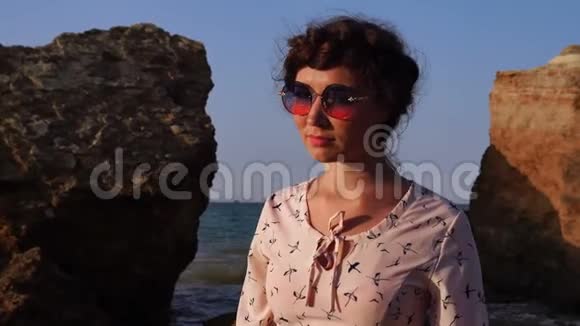 日出时可爱的白种人深褐色的女孩戴着太阳镜和浅粉色的连衣裙坐在石头上视频的预览图