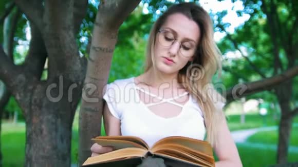 戴眼镜的漂亮年轻女子倚在树枝上看书视频的预览图