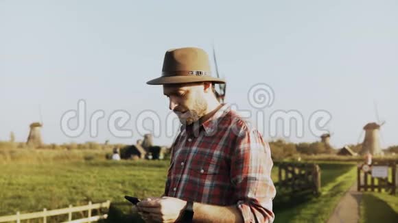 4K成功的欧洲农民男性肖像男人穿着衬衫和帽子带着智能手机和智能手表走向磨坊视频的预览图