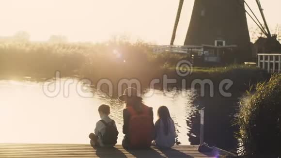日落时有两个孩子的女人坐在湖边码头上令人惊叹的一家人在水边4K后视图视频的预览图
