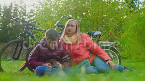 十几岁的男孩坐在草地上玩智能手机坐在妈妈旁边视频的预览图