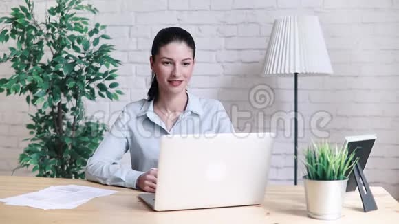 上班时坐在桌旁的一个手提电脑视频通话时微笑的时髦女孩在说话挥手打招呼视频的预览图