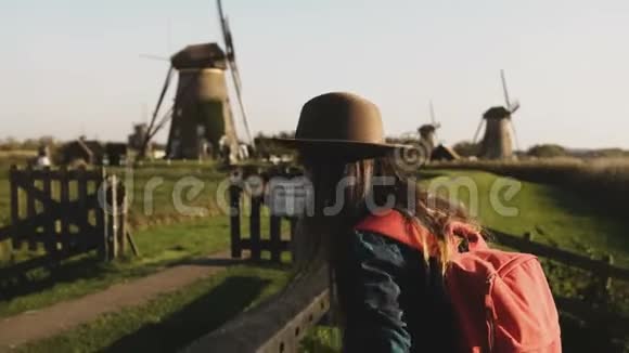 偶然的女孩跳过风车农场附近的篱笆戴着长发和红色背包的女人在日落时回家4K视频的预览图