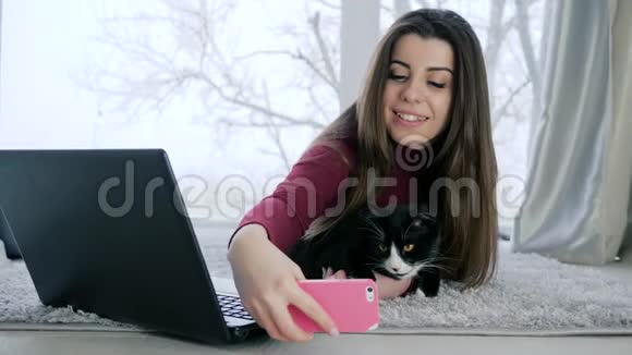 戴着猫的自拍长发女孩在智能手机上被拍到宠物躺在窗户的背景下的地板上视频的预览图