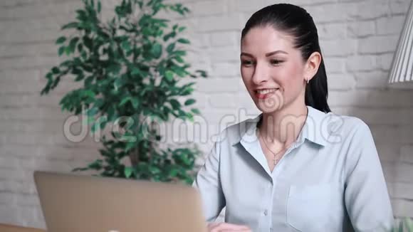 上班时一个手提电脑坐在桌子旁的视频通话中微笑的时髦女孩在聊天挥手和打招呼视频的预览图