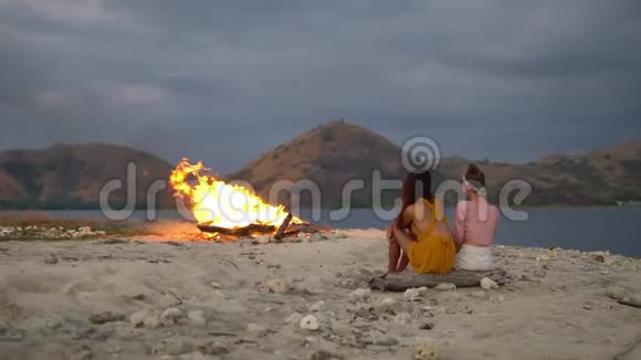 傍晚在海湾的海滩上火在燃烧青山在远处两个朋友坐在一根木头上梦见自己视频的预览图