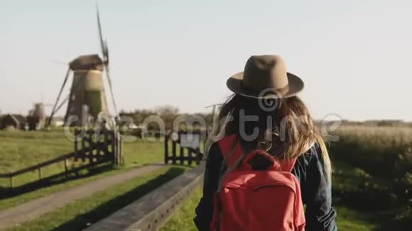 当地女孩慢慢地走到一个风车农场戴帽子的女人留着长发和红色背包回家4K视频的预览图