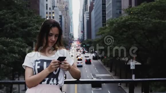 年轻漂亮的女人站在交通道路附近的桥上在美国纽约使用智能手机视频的预览图