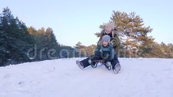 在冬季森林的背景下从雪山上开着大嘴的母亲和儿子乘坐雪橇视频的预览图