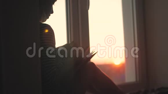 一个小女孩在日落的背景下在窗台上打开的窗户上用书想象故事儿童读物视频的预览图