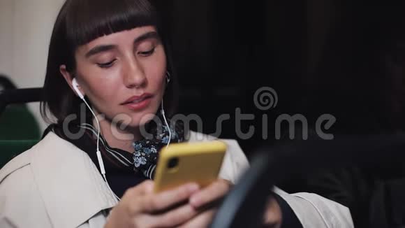 年轻时尚女性戴着耳机听音乐和在公共交通工具上浏览手机的肖像青年视频的预览图