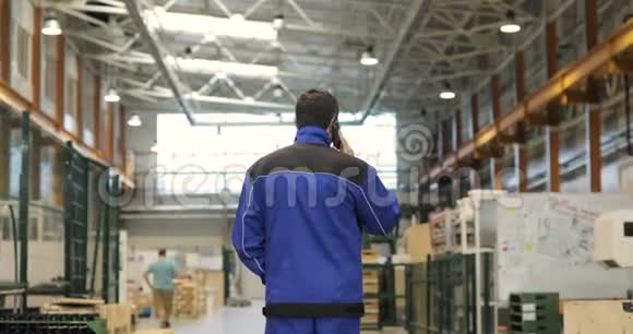 穿着蓝色制服的工程师正在穿过工厂在手机上交谈背面视图视频的预览图