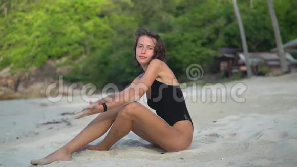 美丽的晒黑了的年轻女子穿着黑色泳衣坐在沙滩上双手放在膝盖上玩沙子视频的预览图